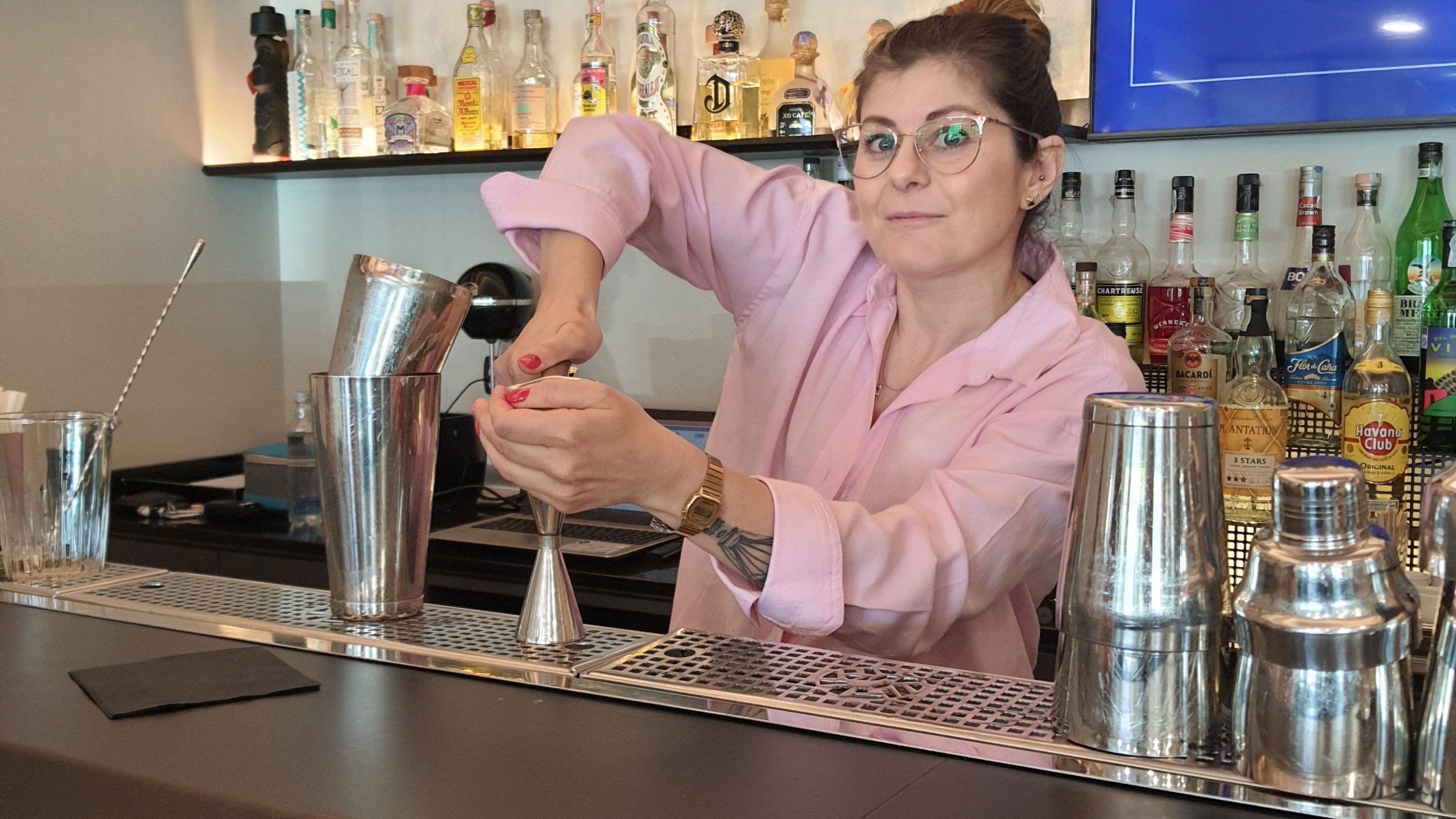 Iulia che sta preparando un cocktail sul bancone di un bar, davanti da una bottigliera piena di bottiglie di alcolici pregiati