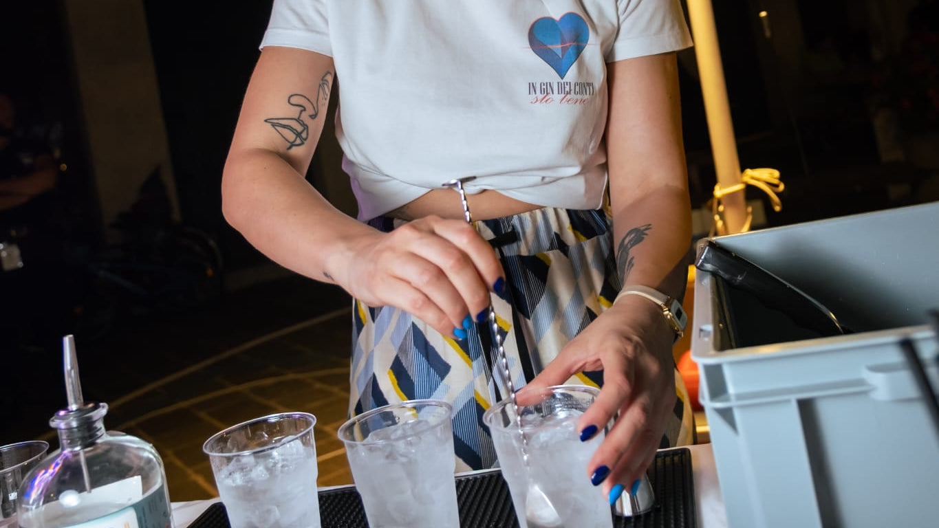 Iulia che sta preparando 3 bicchieri di gin tonic pieni di ghiaccio in primo piano, sta mescolando con il bar spoon. Si vede la scritta "In gin dei conti sto bene" sotto al logo del brand (il cuore blu) sopra la maglietta bianca che indossa Iulia.